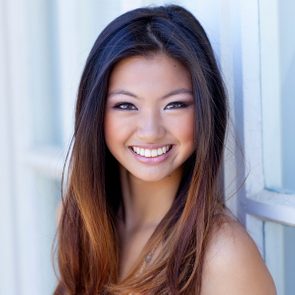 A person smiles warmly, displaying long hair and vibrant eyes, standing against a light-colored wall in a bright, softly lit setting.