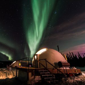Domed structure sits beneath vibrant green aurora, surrounded by snowy landscape and silhouetted trees under a starry night sky.