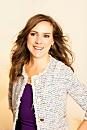 A person smiles, standing confidently, wearing a white patterned jacket over a purple top against a light, neutral background.