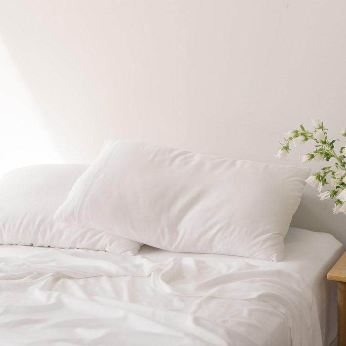 White pillow resting on a bed, with white sheets slightly wrinkled; a vase of white flowers sits on a bedside table nearby.