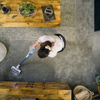 Person vacuuming carpet in a cozy living room with wooden furniture and decorative plants, viewed from above.