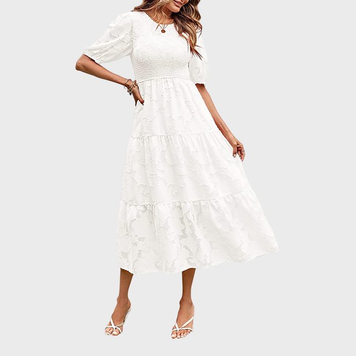 Woman wearing a flowing white dress with puff sleeves, standing in an elegant pose, against a plain light gray background.