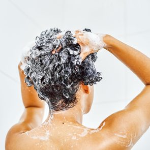 Person washing hair, hands lathering shampoo, in a tiled shower.