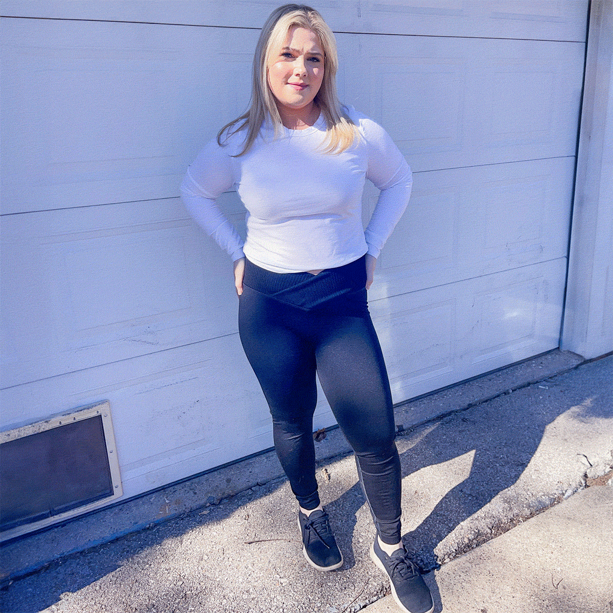 Person standing confidently, hands in pockets, in front of a white garage door. They're wearing a white long-sleeve top and black leggings.