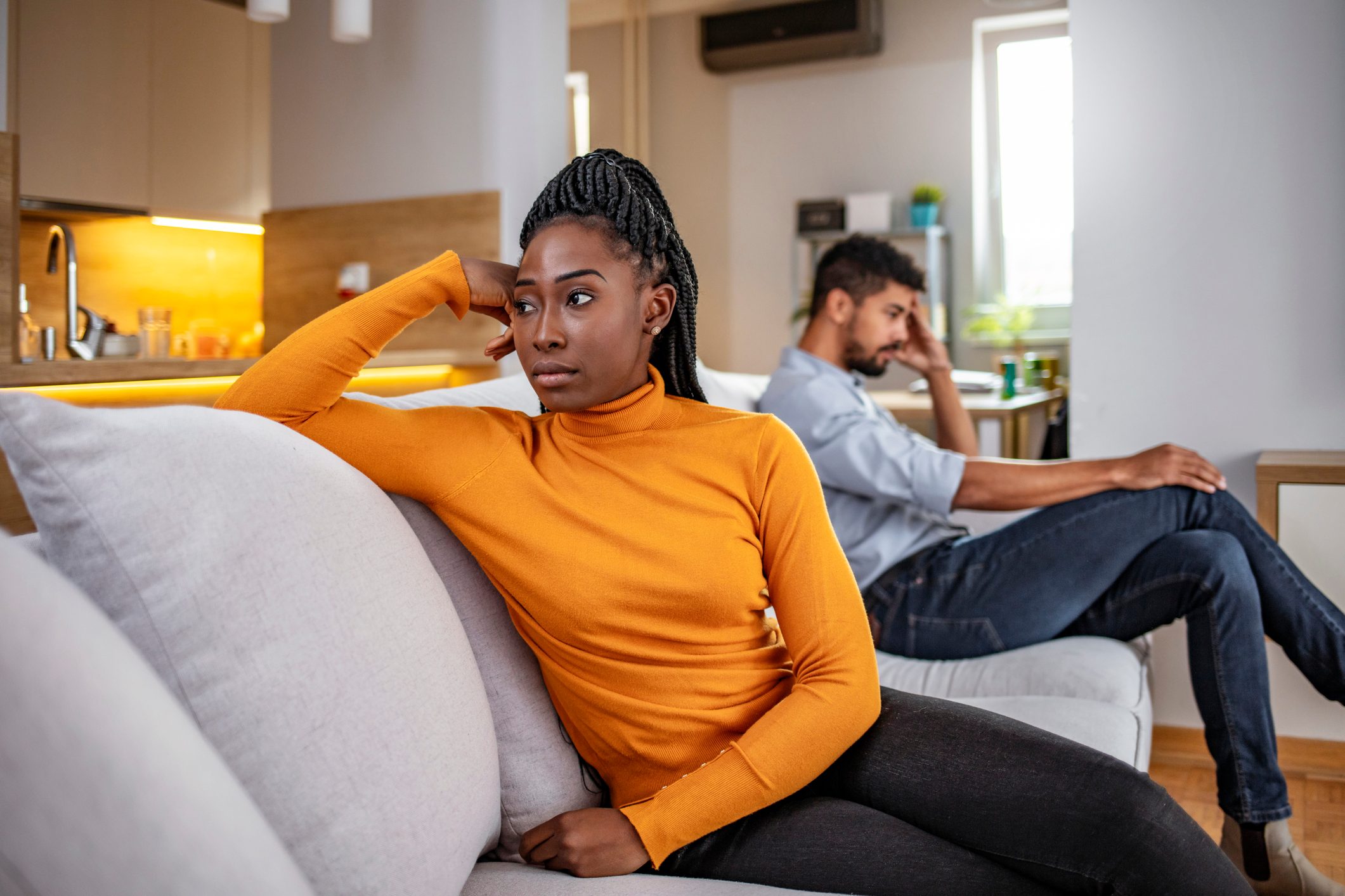 Two people sit on a couch, looking in opposite directions, appearing thoughtful. The setting is a modern living room with kitchen in the background.