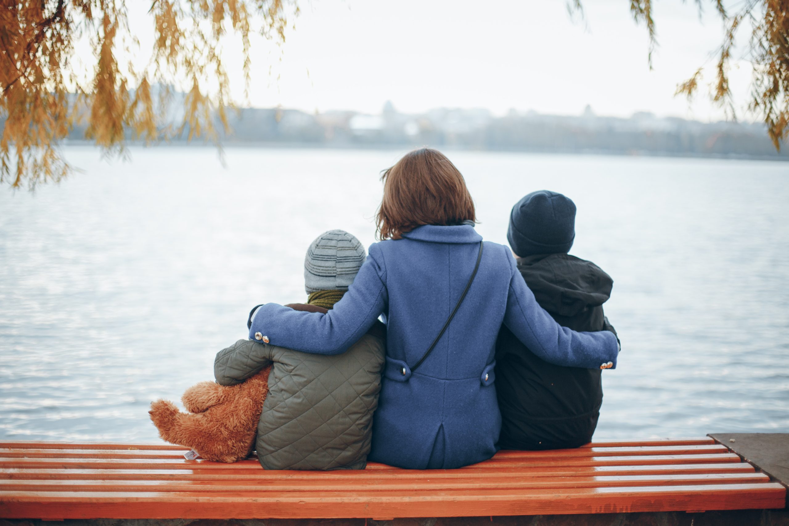 Three people sit on a bench, hugging and facing a calm lake, surrounded by autumn trees.