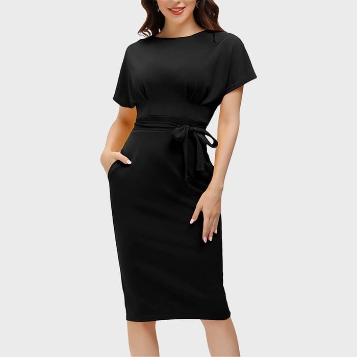 A woman stands wearing a black knee-length dress with a belt, hands in pockets, against a plain gray background.