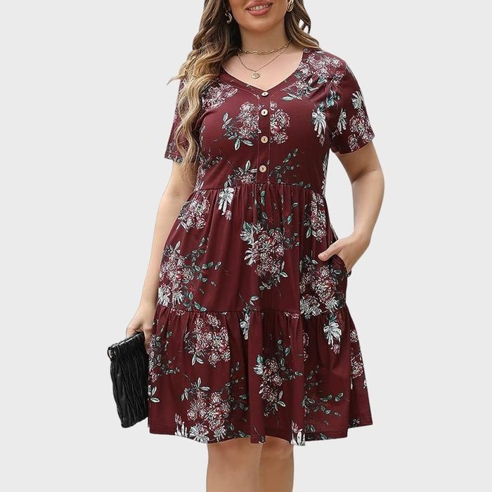 Woman wearing floral dress, holding black clutch bag, smiling; neutral gray background.