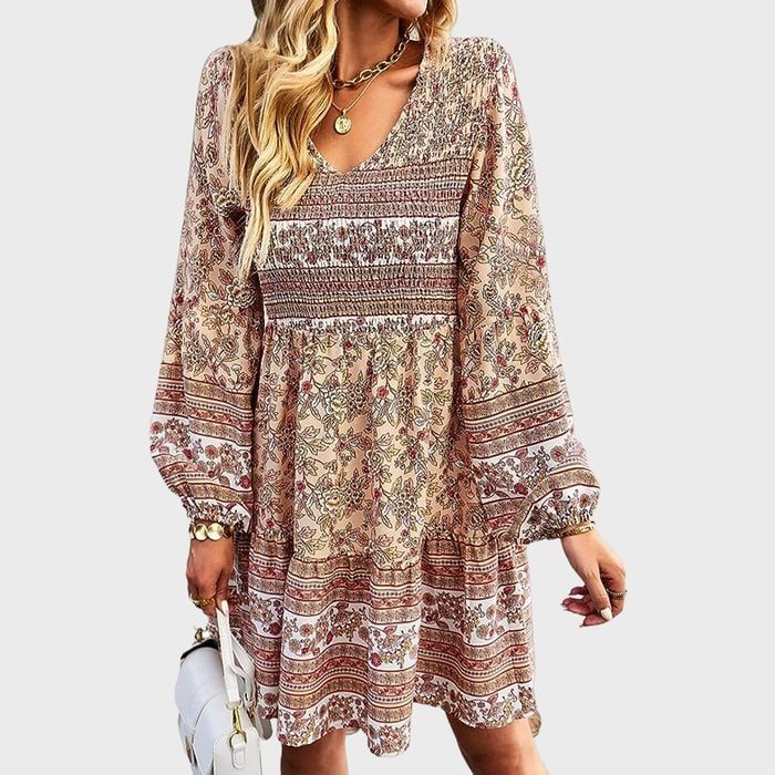 A woman wears a bohemian-style, floral-patterned dress, holding a white handbag in a neutral background.