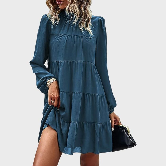 Woman wearing a flowing, long-sleeved teal dress holds a black handbag. Background is light gray.