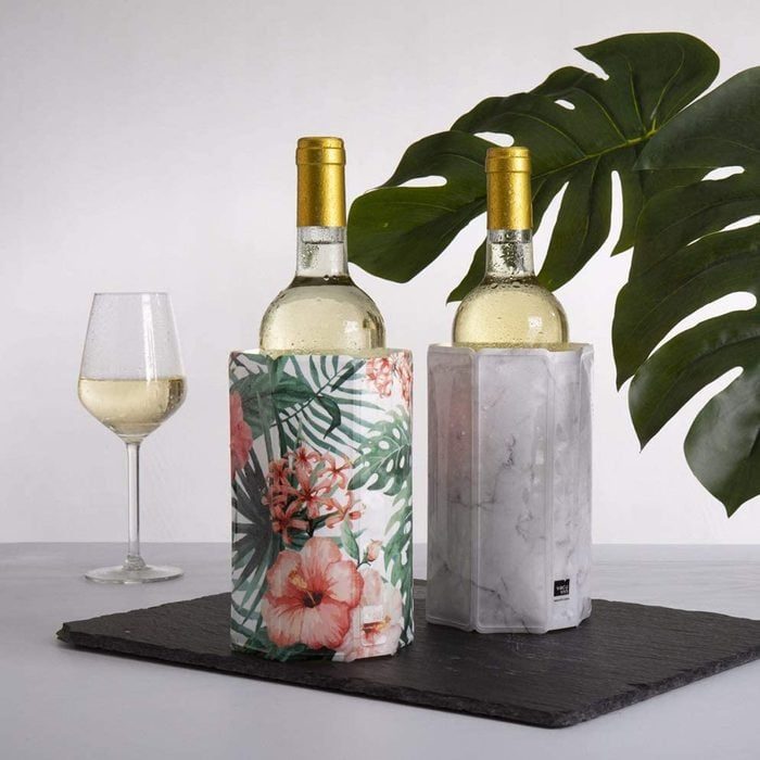 Two wine bottles sit in decorative sleeves on a slate board, accompanied by a filled wine glass, with a large leaf in the background.