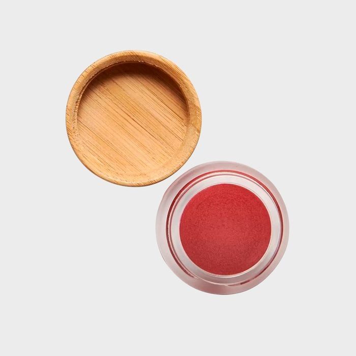 Red cosmetic balm sits in an open glass container next to a circular wooden lid on a plain light gray background.