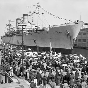 A large crowd gathers at a dock to greet a ship covered with flags and banners. The ship is moored at the pier, and people are holding signs as they look toward the vessel. Buildings are visible in the background.