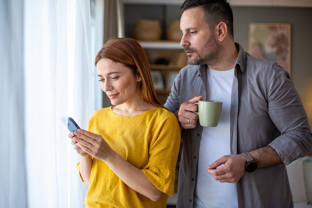 A woman is looking at her phone and a man sneaks up from behind to peek at what she's doing
