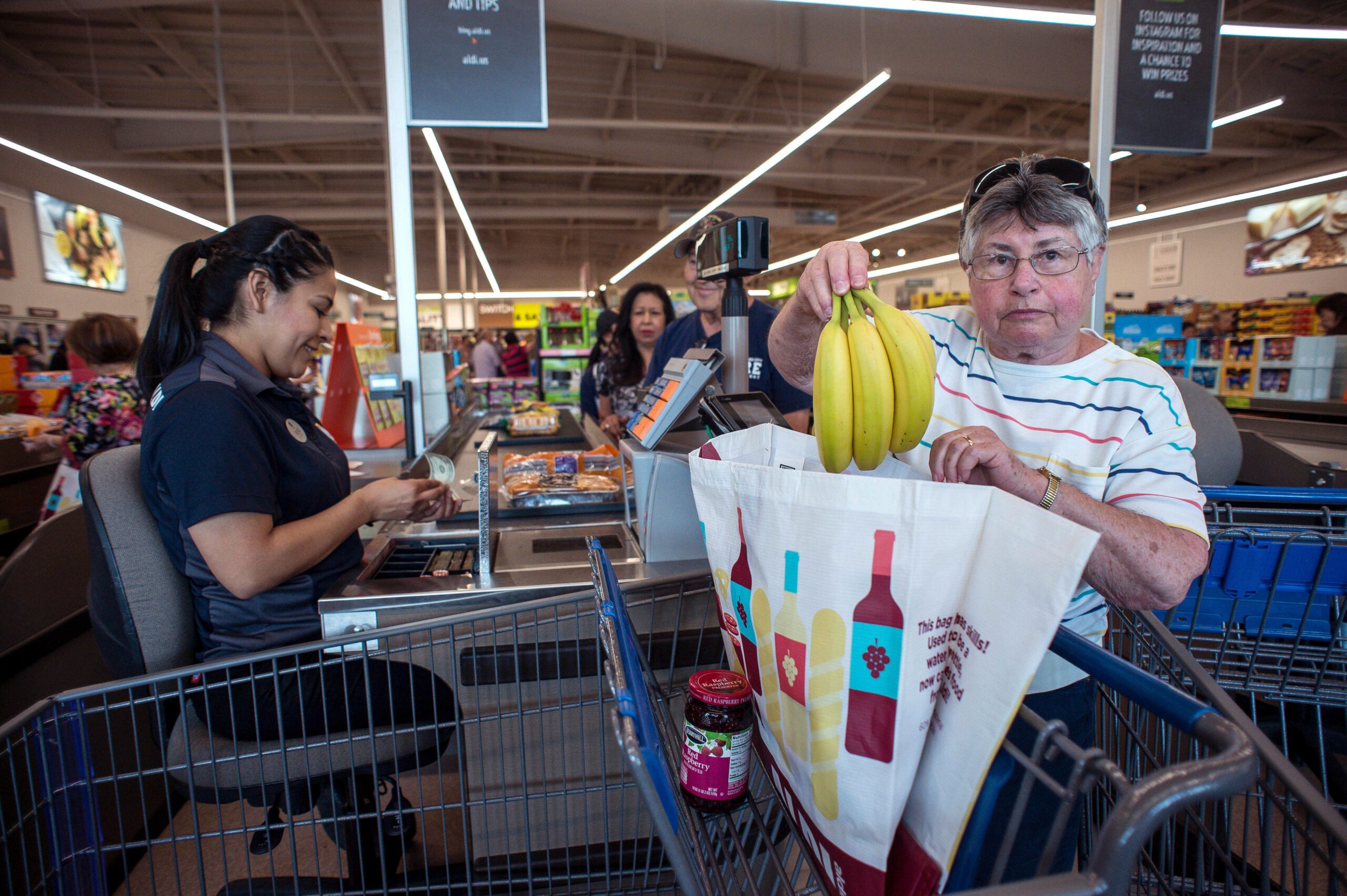Cashier (left) sitting down at an Aldi checkout line while a customer puts bananas into a grocery bag