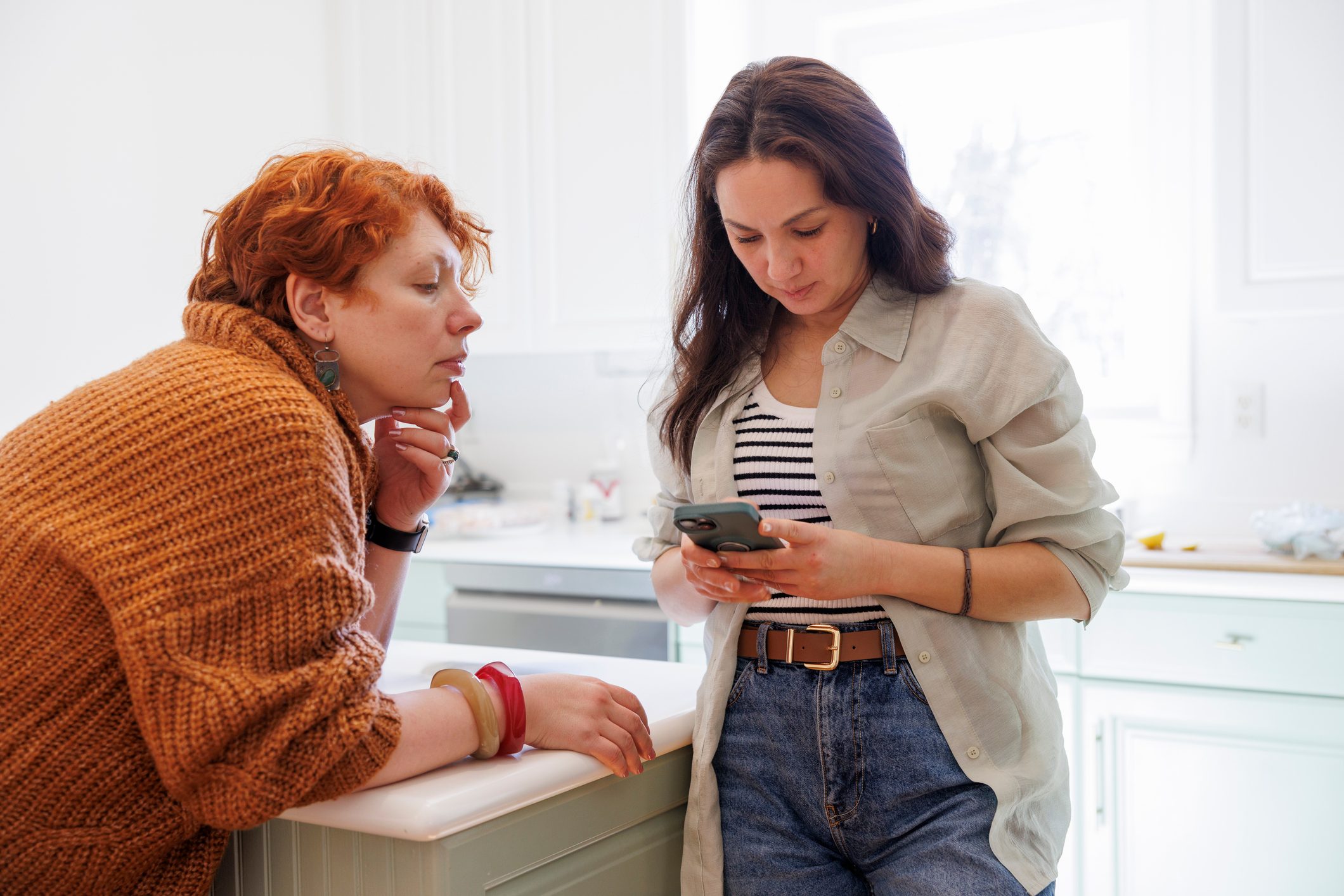 Friends distracted by smartphone. Redhead and Asian women in kitchen during conversation.