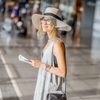 Young woman in grey dress and sunhat standing with suitcase and boarding pass at the departure hall of the airport