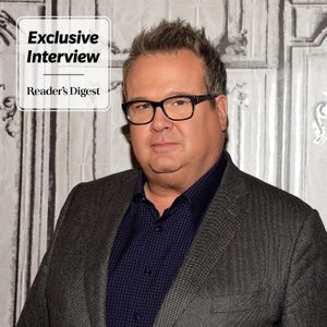 Eric Stonestreet in a blazer on a white wooden background with an 